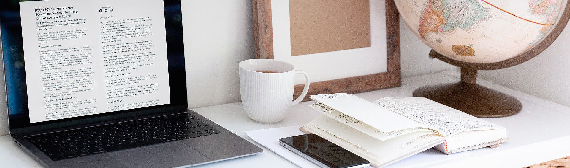 A laptop displaying a document next to a cup, a globe, a notebook, and a framed blank picture on a desk.